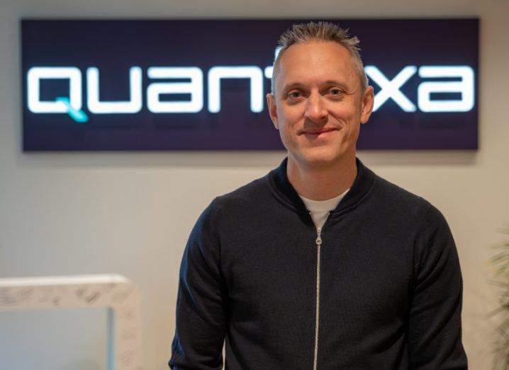 A man wearing a dark zipped up jacket smiles in front of a wall bearing the Quantexa logo. He is Jamie Hutton, Quantexa's chief technology officer.