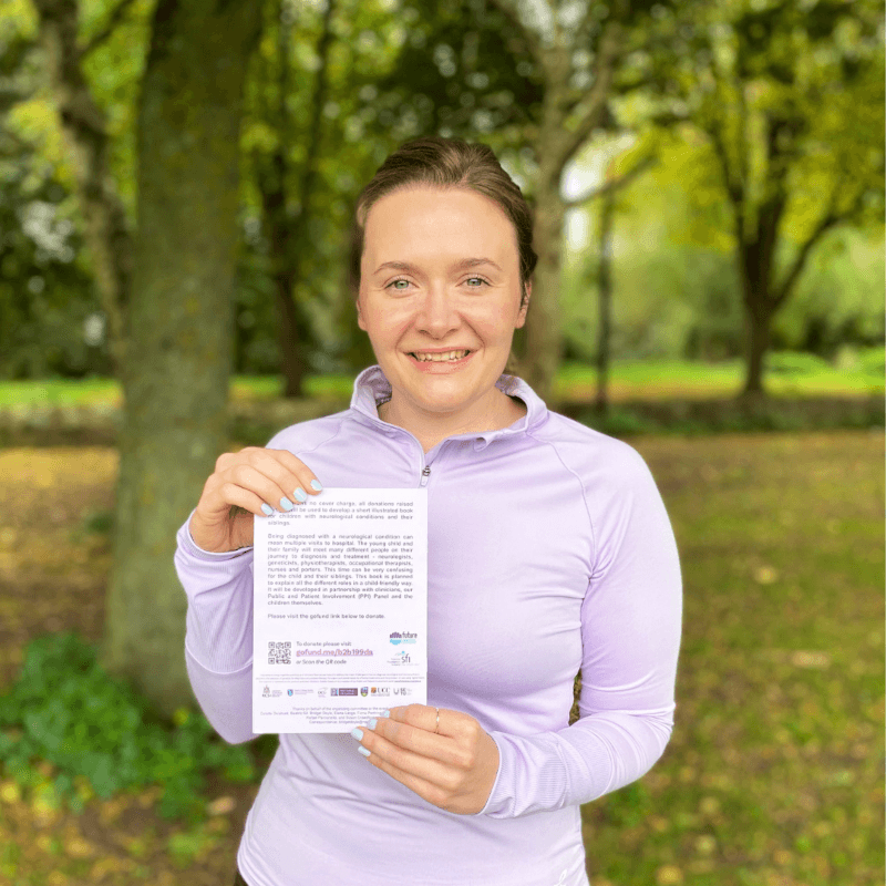 Amy Holtby holds up a QR code for her fundraising effort after running the Dublin city marathon. She wears lavender active wear and stands in a park.