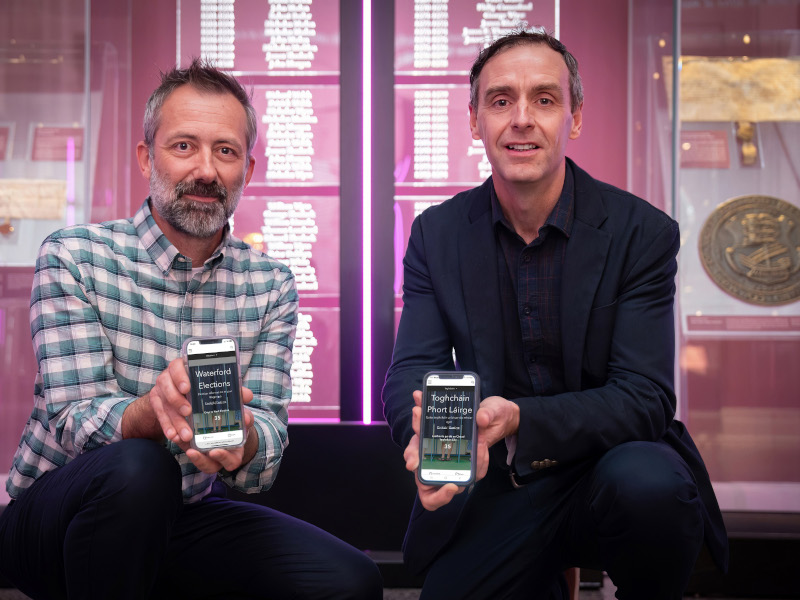 Two men sitting down and holding phones that show the Waterford elections hub.