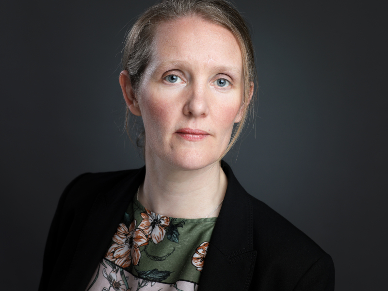 A headshot of Sheelagh Brady who wears a black blazer and a dark shirt with a flower pattern. She has fair hair, worn up and she stands against a grey backdrop.