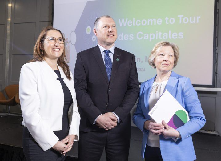 Two women and a man stand in front of a projector screen that reads Tour des Capitales.