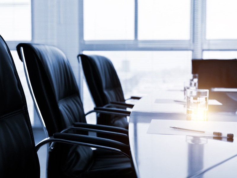 A boardroom close-up of black chairs and white paper, pens and a glass on a table with windows and the sun's rays shining through.