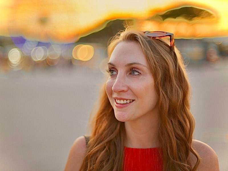 A headshot of Sarah Kerr smiling and looking to the left of the frame. She has long red hair, wears a red top and sunglasses on her head. There is a sunshine evening glow.