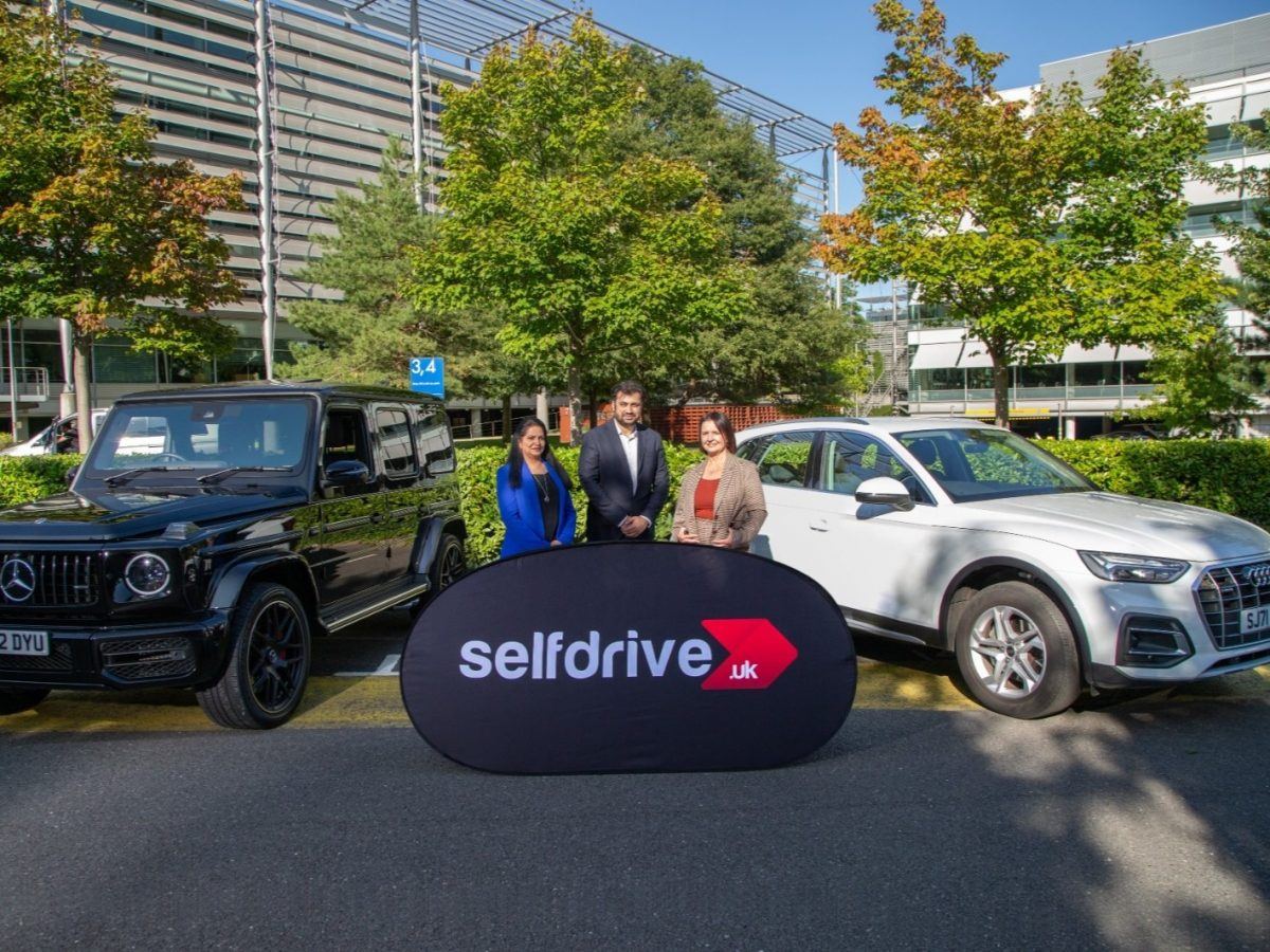 Two women and a man stand behind a sign that has the SelfDrive logo on it.