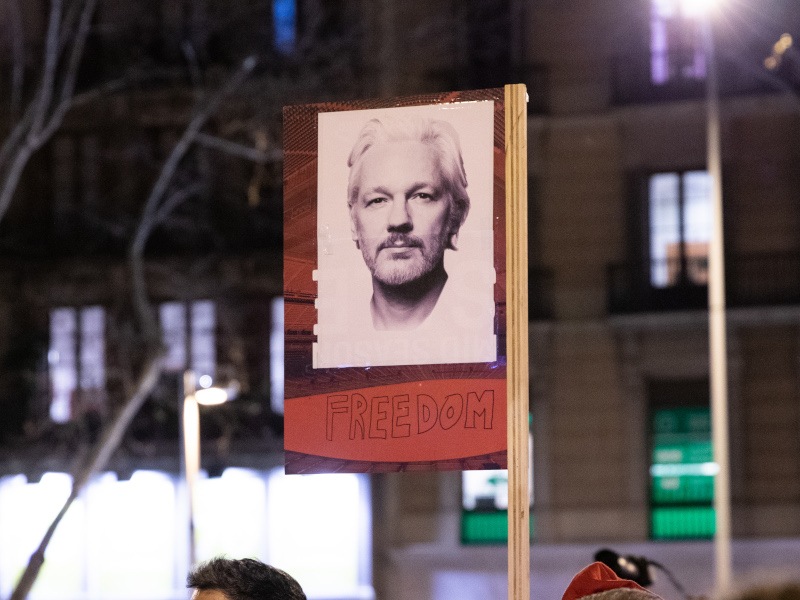 A photo of Julian Assange on a poster during a protest, with buildings in the background.