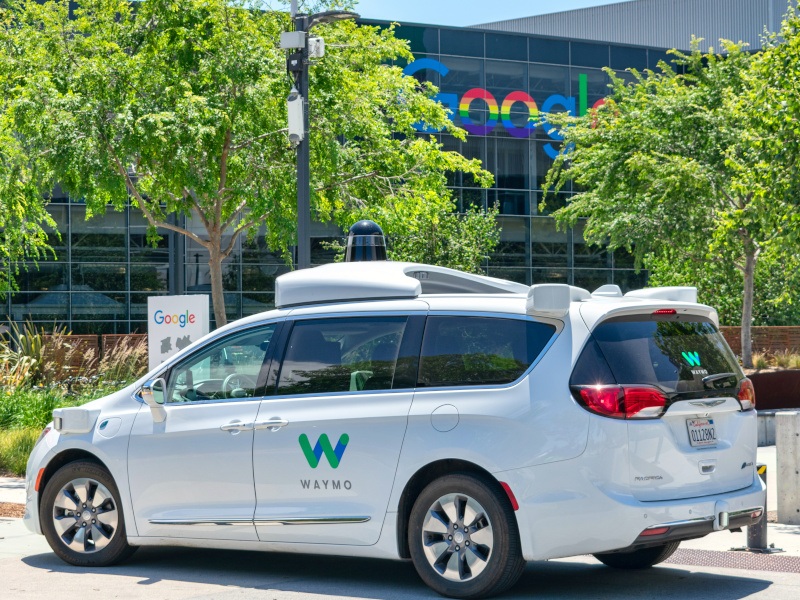 A white vehicle with the Waymo logo on the side, parked in front of a building with the Google logo on the side.