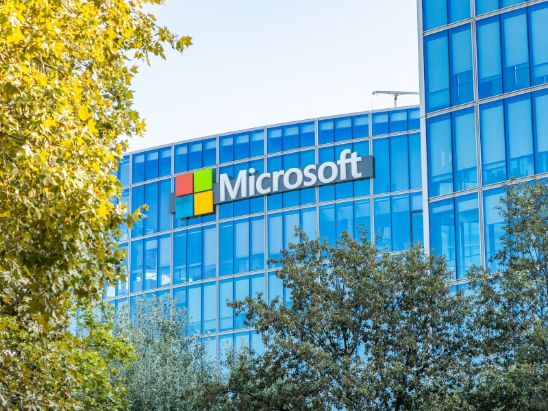 The Microsoft logo on the side of a tall building with trees in the foreground of the image.