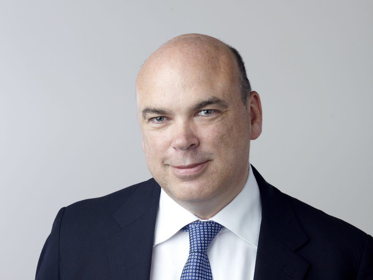 A close-up headshot of Mike Lynch in a dark suit with a white shirt and blue tie, against a grey backdrop.