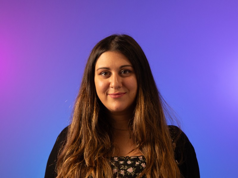 A woman with long brown hair smiles at the camera in front of a purple virtual background.