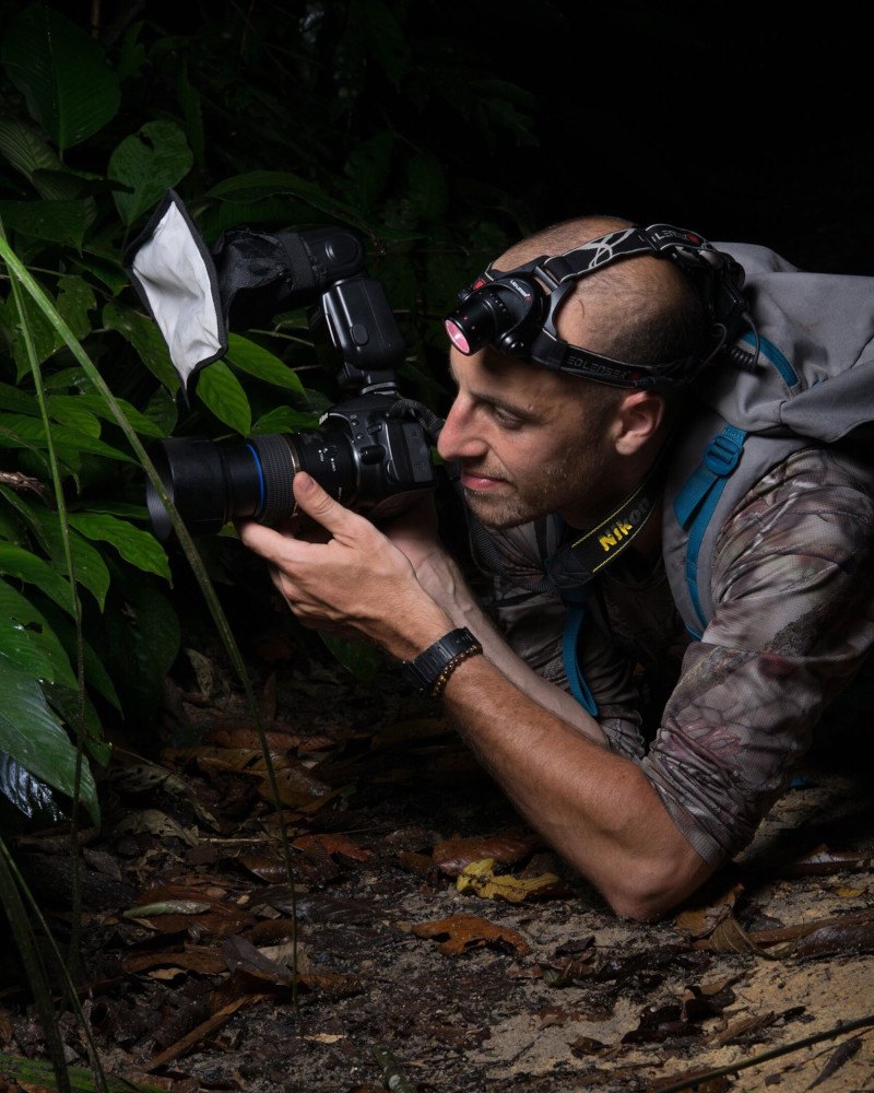 Sami Assad kneeling down in a forest with a camera in his hands and a torch strapped to his head.