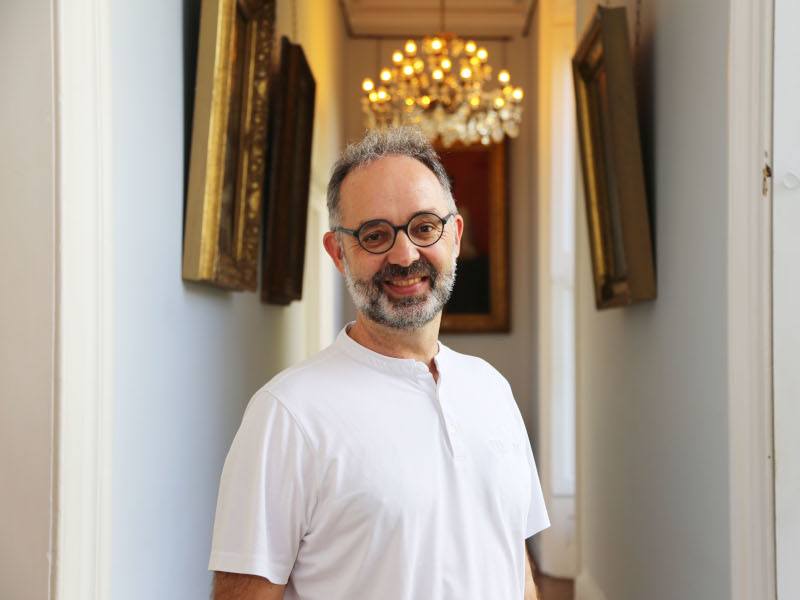 A man wearing a white shirt and glasses smiles in a hallway in front of a chandelier. He is Dr Ruediger Kuehr.