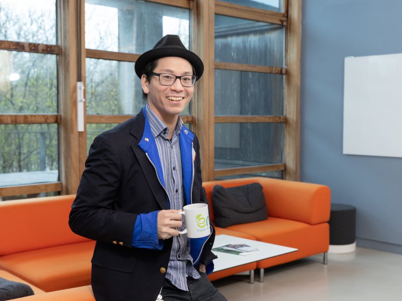 Doctor Kian Mintz-Woo stands smiling wearing a hat and a suit jacket over a blue shirt, holding a mug. There is a bright orange couch and large glass doors and windows behind him.