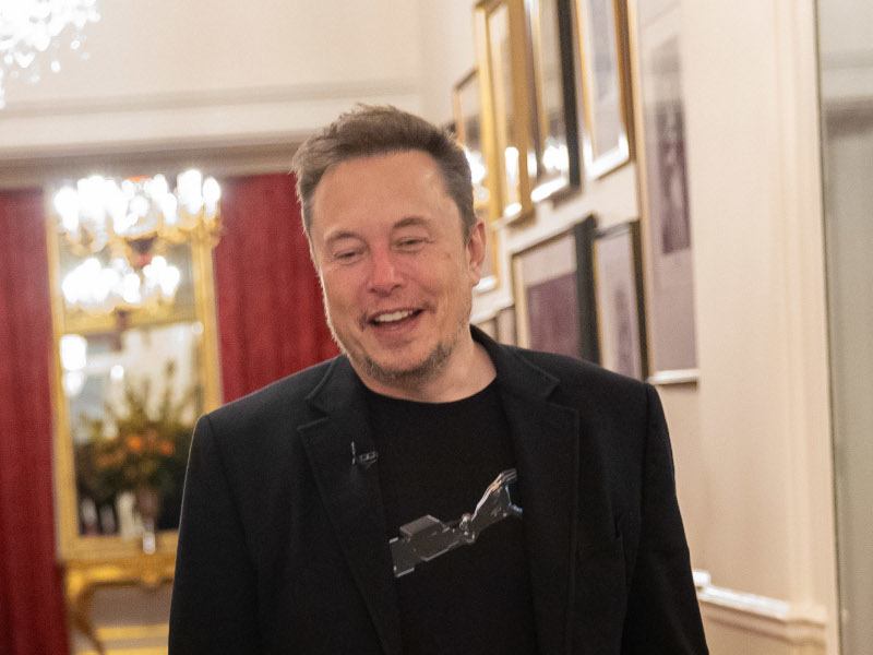 An image of Elon Musk smiling while walking down a hallway, with lights and red curtains in the background.