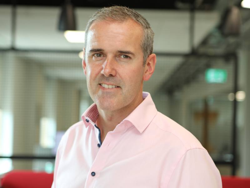 A man in a pink shirt standing in an office, with a red chair and windows behind him. He is Peter Lantry of Equinix Ireland.