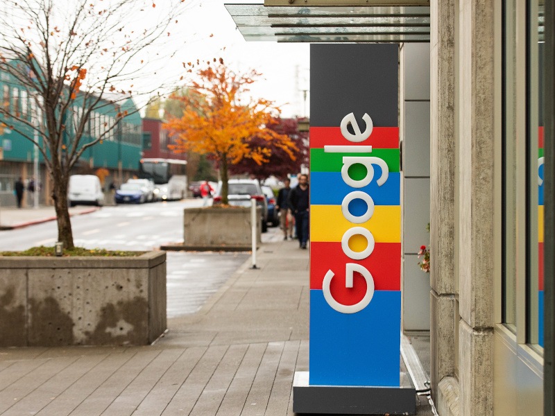 The Google logo on a vertical sign next to a building entrance on a street, with a grey sky in the background.