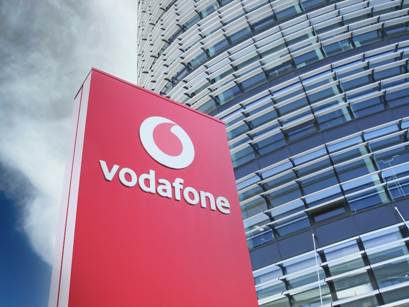 The Vodafone logo on a red sign in front of a tall building, with a cloudy sky overhead.