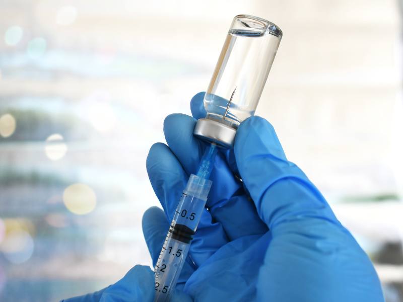 A person's hands in blue gloves, holding a syringe into a vial that contains a clear fluid.