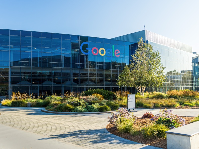 The Google logo on the side of a large building, with trees and smaller plants in front of the building and a blue clear sky above.