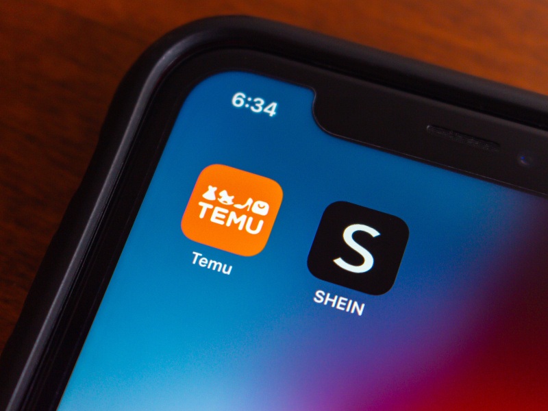The Temu and Shein app logos on a smartphone screen, which is laying on a wooden surface.