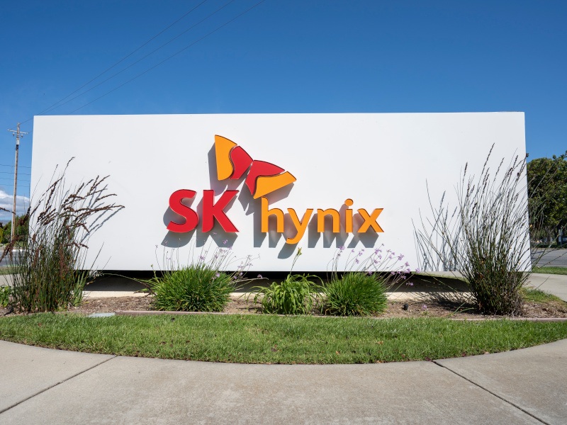 The SK Hynix logo on the front of a white building, with green bushes in front of the building.