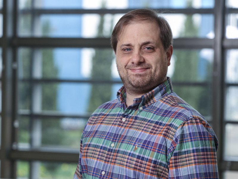 Denis Collins stands slightly turned but facing the camera and smiling in front of a window with slats on it and greenery visible outside. He wears a blue and red check shirt.