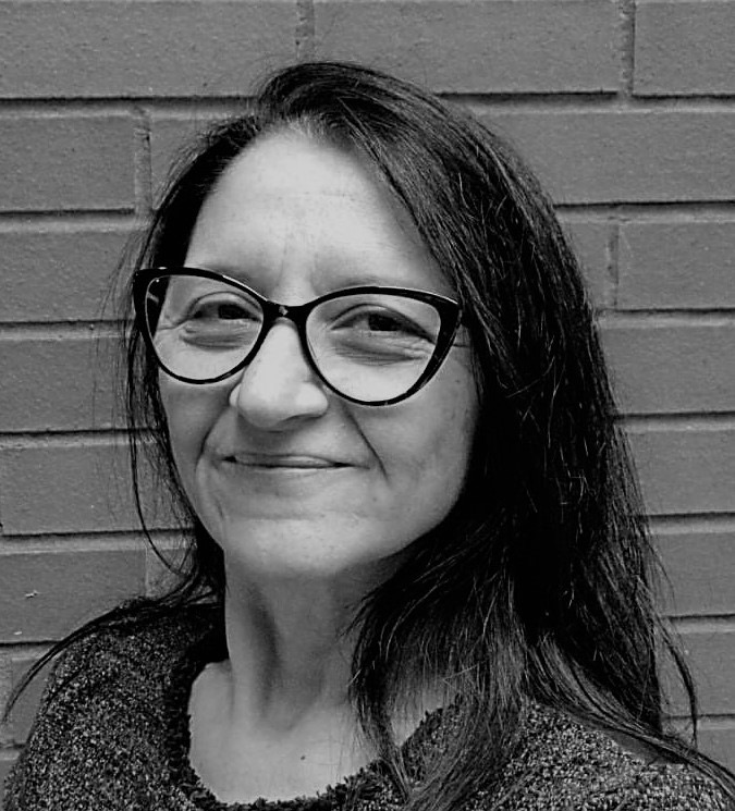 A black-and-white headshot of Dr Maria Aretoulaki. She wears glasses and has long dark hair and smiles at the camera in front of a brick wall.
