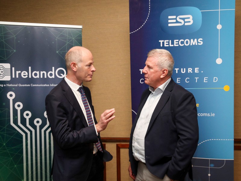Two men speaking in front of two banners, which show the ESB Telecoms and the IrelandQCI logos.