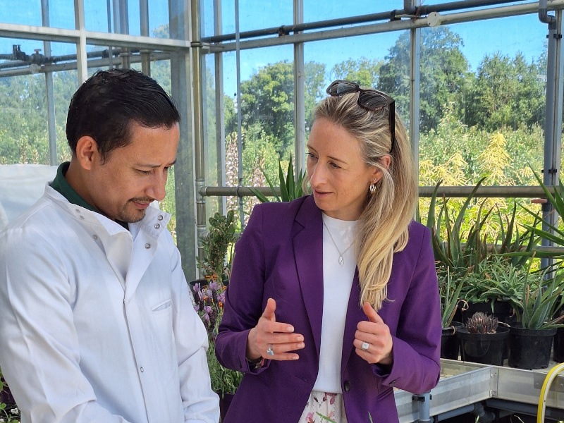A man in a lab coat stands with a woman with blonde hair wearing a purple coat. They are standing in a greenhouse on a sunny day.
