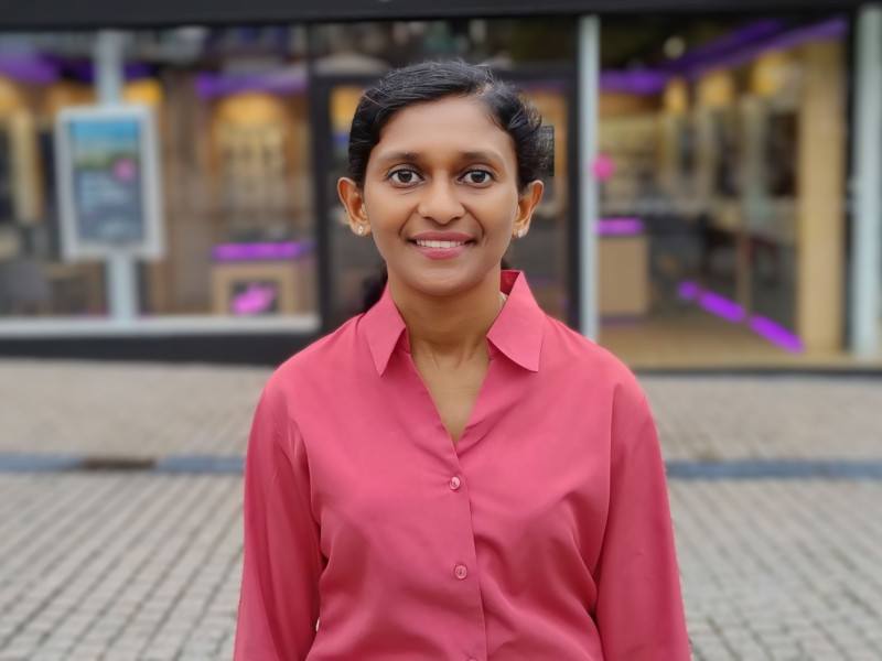 Dr Lizy Abraham stands outside on a paved road in front of a shop window. She wears a pink shirt and smiles at the camera.