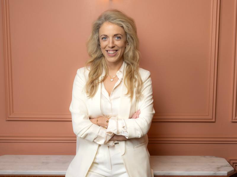 CEO of Platform94 Marie Donnellan poses in a white pants suit against a peach coloured wall.