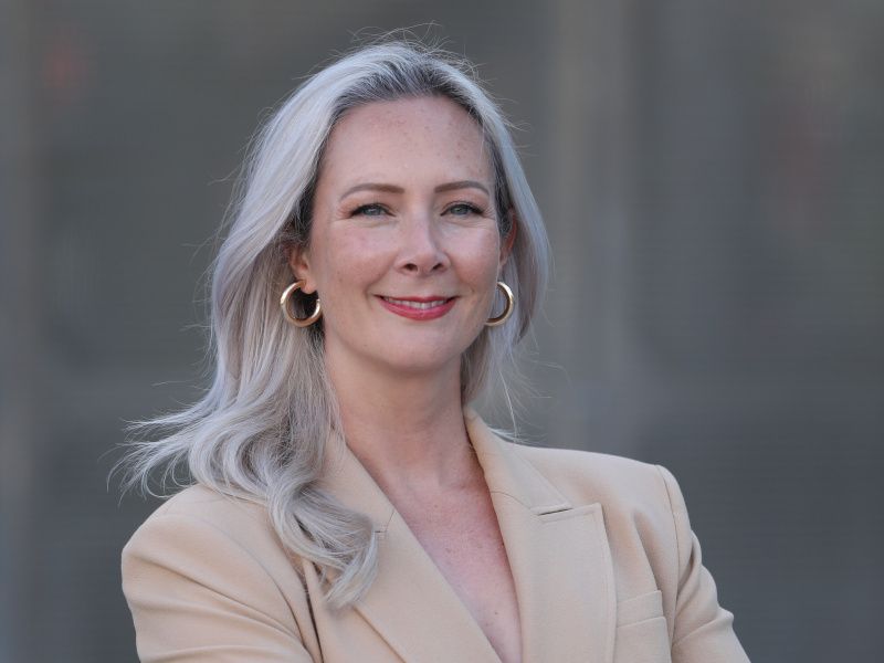 A close-up of a woman with long, grey hair and a beige blazer who smiles at the camera.