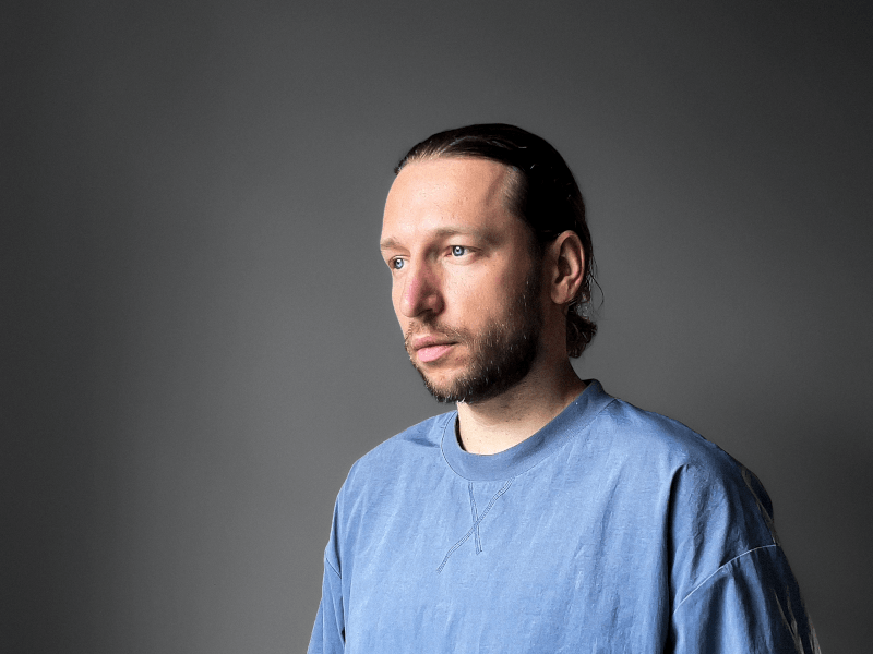 A close-up of Oleksii Fedorenko. He wears a blue t-shirt and looks to the left of the camera, standing against a dark grey background.