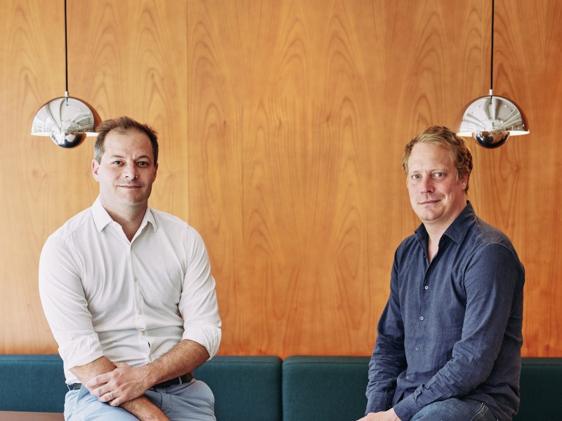 Photo of two men, both co-founders of Qover, sitting on a green couch in front of a wood-panelled wall..