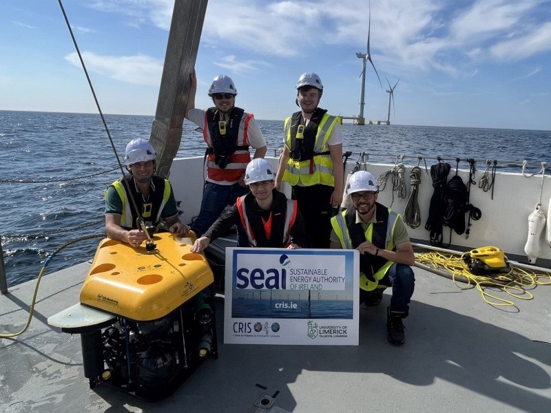 A group of men in high visibility vests standing next to a robotic vehicle, with offshore wind turbines in the background.