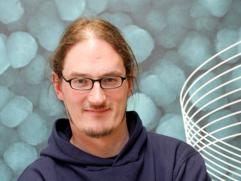 A headshot of Jonathan Coleman. He has brown hair and wears glasses and a navy hoodie. He stands in front a a wall with blue circles on it.