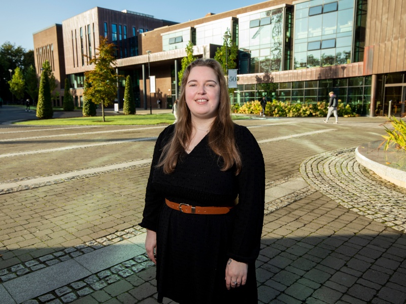 Sarah Guerin stands in the centre of the University of Limerick campus with a square and buildings behind her. She wears a black dress and has long brown hair.