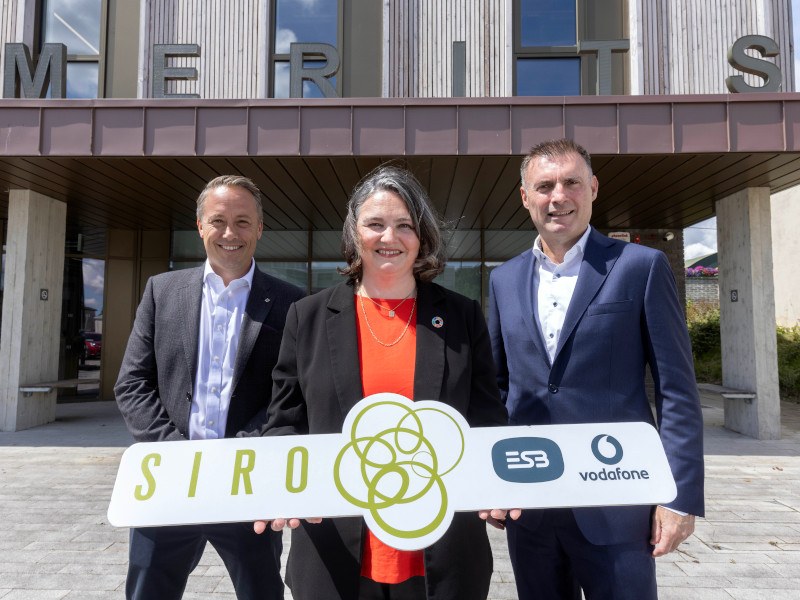 Two men and a woman standing in front of the Merits building. The woman is holding a sign with the Siro, ESB and Vodafone logos on it.