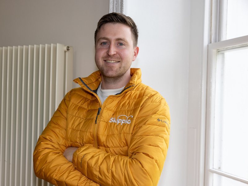 A young man with dark hair stands with arms crossed smiling at the camera. He is wearing a yellow puffer jacket with the Skippio logo on it.