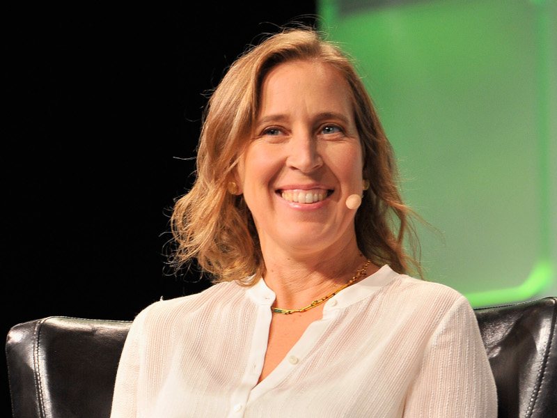 An image of Susan Wojcicki, smiling while sitting on a chair with a small microphone connected to her ear.