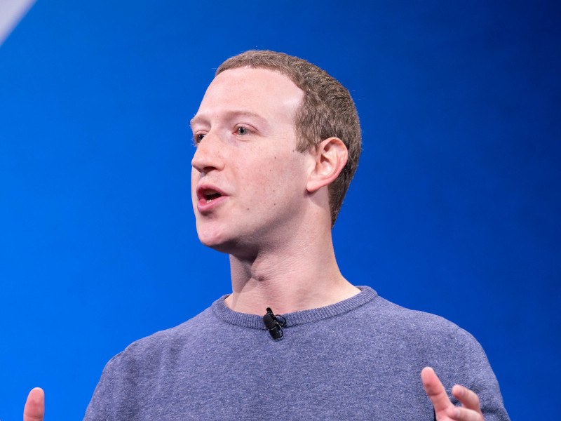 Mark Zuckerberg standing on a stage and speaking, with a blue wall behind him.