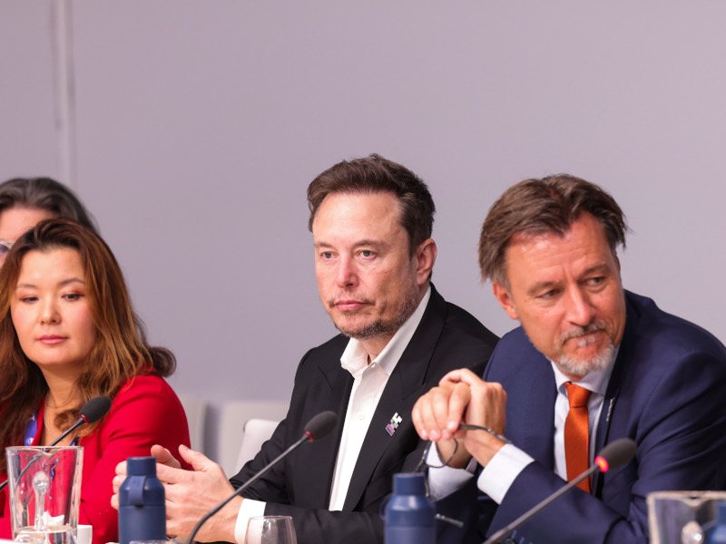 Elon Musk sitting next to a man and a woman with a microphone in front of him.