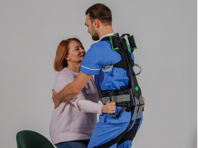 An image of a male healthcare worker wearing an exoskeleton while helping a woman stand up from a chair.