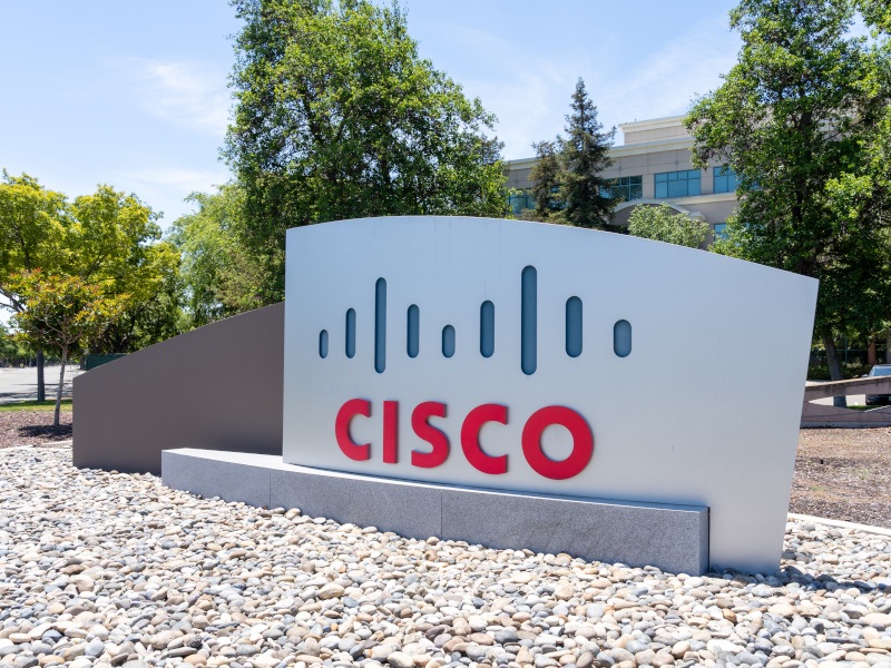 The Cisco logo on a sign in front of multiple trees and a building.