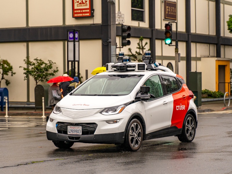 A Cruise robotaxi driving in the middle of a road with a traffic light and a building behind it.