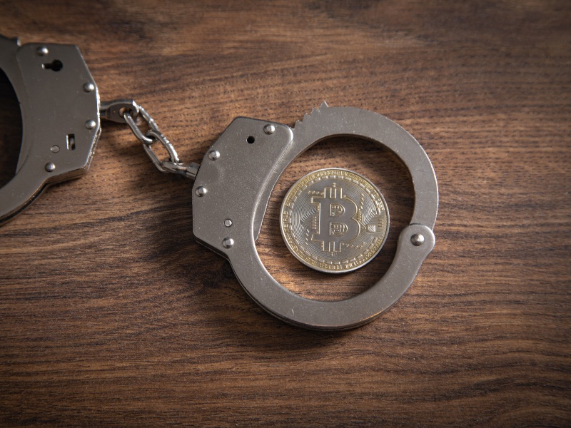 A pair of handcuffs with a coin showing the Bitcoin logo in one of the cuffs, on a wooden table.