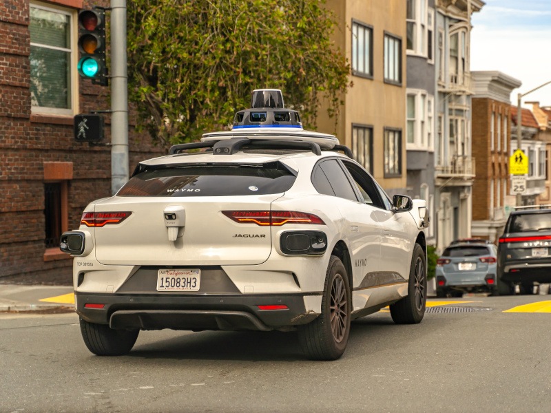 A Waymo vehicle driving behind other cars on a road, with a green traffic light and buildings in front of it.