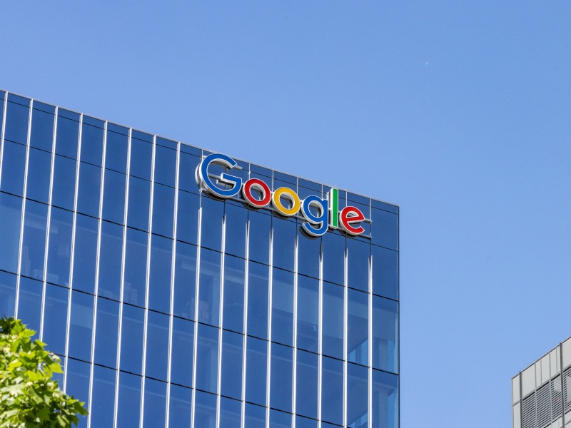 The Google logo on the side of a tall building near the roof, with a clear blue sky behind it.