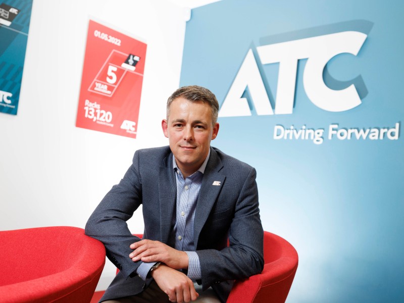 Keith Young, MD of ATC, sits in a red chair with his company logo on a blue wall behind him.