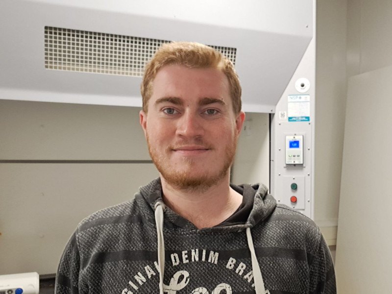 A headshot of Ciarán Cooling. He wears a grey hoodie and stands in a lab.