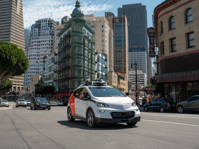 A Cruise autonomous vehicle driving down a road with cars and buildings behind it.